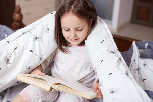 Horizontal indoor shot of playful smart sweet kid holding book in both hands, looking through attentively, sitting in her bedroom under blanket, wearing pyjamas. Children and free time concept. — Stock Photo, Image