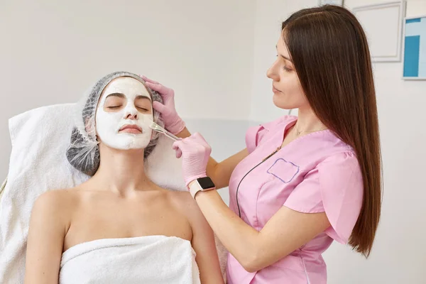 Poziome zdjęcie pomocnego, ciężko pracującego kosmetologa nakładającego białą maskę na skórę profesjonalną szczotką. Rozkoszne leżenie z zamkniętymi oczami, zabiegi piękności. Koncepcja leczenia skóry. — Zdjęcie stockowe