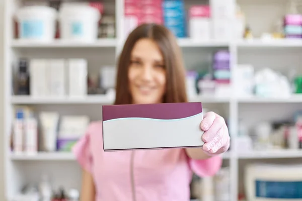 Esteticista alegre positivo em pé em seu estúdio sozinho, sorrindo sinceramente, segurando pacote cosmetológico em primeiro plano, estando em fundo turvo, tendo muitas prateleiras à tona. Conceito de beleza . — Fotografia de Stock