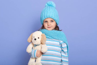Stüdyoda elinde oyuncak köpekle mavi duvara yaslanmış, çizgili gömlek, ponponlu eşarp ve şapka takmış, doğrudan kameraya bakan, siyah saçlı, oyuncağıyla oynayan küçük bir kız görülüyor..