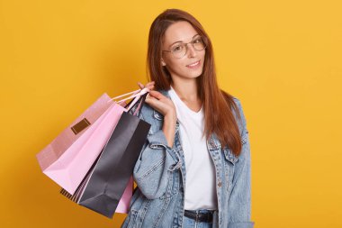 Sarı stüdyo arkaplanından izole edilmiş pembe ve siyah alışveriş çantaları taşıyan düz saçlı güzel bir kadının portresi, doğrudan kameraya bakıyor, günlük giysiler ve gözlük takıyor..