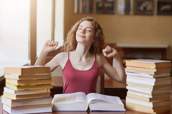 Beautiful female student in university library siiting at table with fists up, keeping eyes closed, sits in front opened book, woman surrounded with books, prepares for examinations, being tired. — Stock Photo, Image