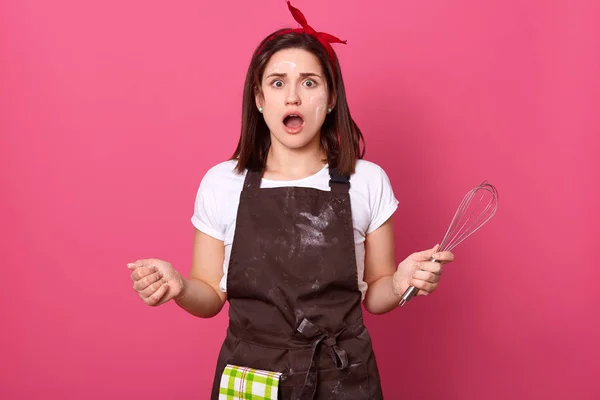 Ama de casa mujer cocinero panadero vestidos delantal marrón, camiseta blanca y banda roja de la cabeza posando aislado sobre fondo de pared rosa. Impactada ama de llaves loca extendiendo la mano, mostrando gesto indefenso . — Foto de Stock