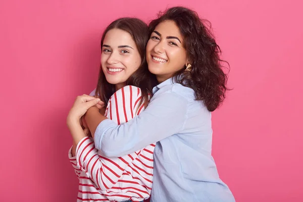 Portrait of caucasian young women isolated over rosy background, stand smiling, wavy haired lady embracing her friends who stands backwards to her, girls expressing happyness. One sex love concept. — 스톡 사진