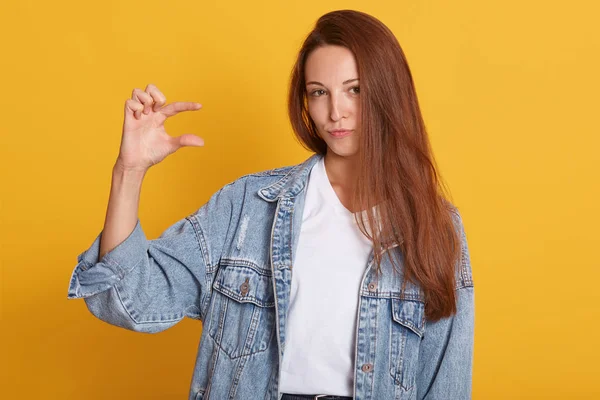 Mooie vrouw met straighet donker haar dragen denim shirt en casual wit shirt poseren over geïsoleerde gele achtergrond, gebaren met de hand, tonen kleine formaat bord met vingers, kijken naar camera — Stockfoto