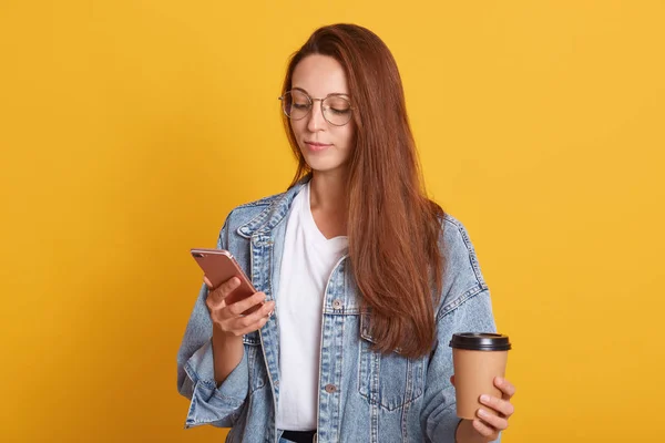 Studio shot dari stupefied Caucasian woman reading news on internet website while drinking takeaway coffee, holding mobile phone, dress white shirt and denim jacket, looks calm, poses indoor . — Stok Foto