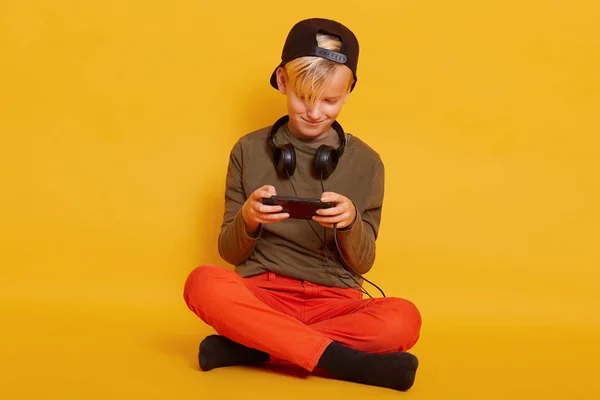 Retrato de cerca del pequeño rubio con ropa casual, posando con auriculares alrededor del cuello, jugando videojuegos en línea a través del teléfono móvil, se ve concentrado, aislado en el fondo amarillo . — Foto de Stock