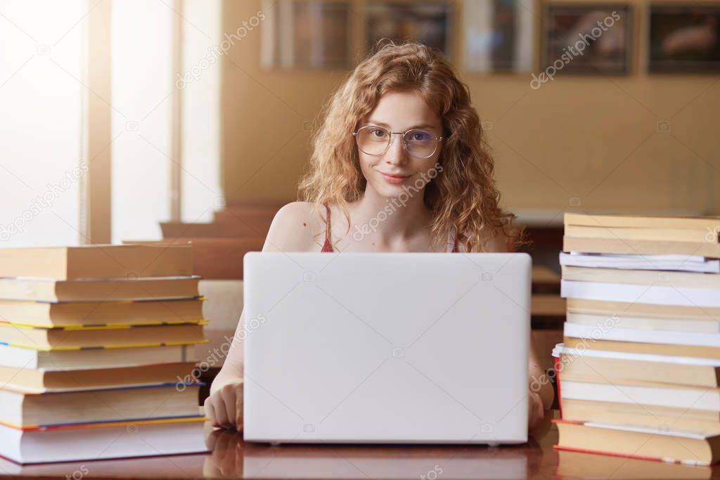Close up portait of teenage girl studying and using new technologies, sitting at table in front of opened white lap top, lady wears t shirt, student surrounded with different books. Education concept.