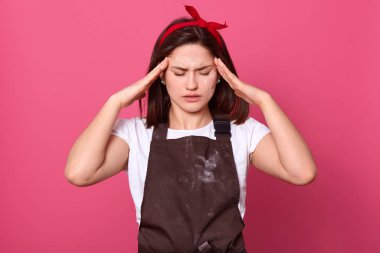 Depresyonda olan, siyah saçlı, başı ağrıyan, elleri şakaklara koyan, gözlerini kapatan, yorgun, günlük giysiler giyen, ev işleri yapan genç bir kadının fotoğrafı..