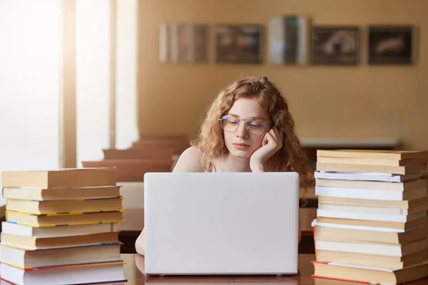 Horizontal picture of cute beautiful charming attentive female looking at laptop screen, wearing eyeglasses, using modern device, studying hard, spending time alone. People and studying concept. — Stock Photo, Image