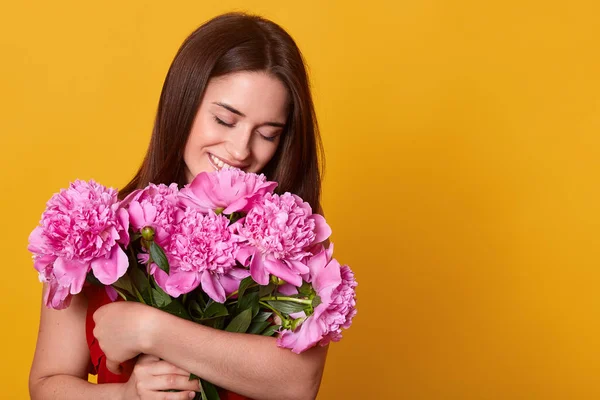 Indoor studio shot of tender charming young female having pleasant facial expression, smelling pink peonies, getting pleasure of presents, being in high spirits. Copyspace for advertisement. — 스톡 사진