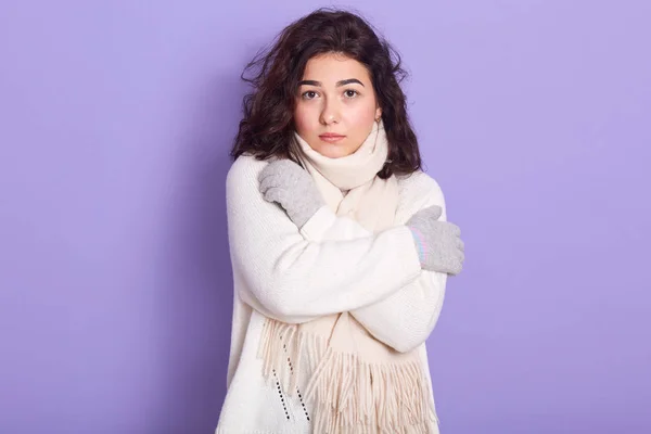 Retrato de mujer atractiva seria con el pelo negro rizado mirando directamente a la cámara, cruzando brazos, usando suéter blanco, bufanda y guantes grises, de pie aislado sobre fondo lila en el estudio . — Foto de Stock