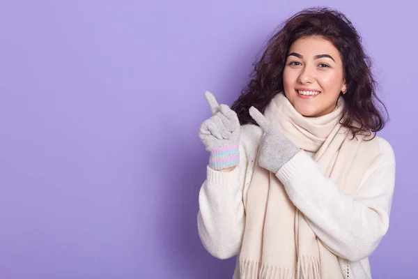 Retrato de cerca de una joven alegre y guapa con el pelo oscuro rizado, pasando tiempo a solas, haciendo un gesto con el dedo índice, usando suéter blanco, bufanda y guantes. Copyspace para publicidad . —  Fotos de Stock