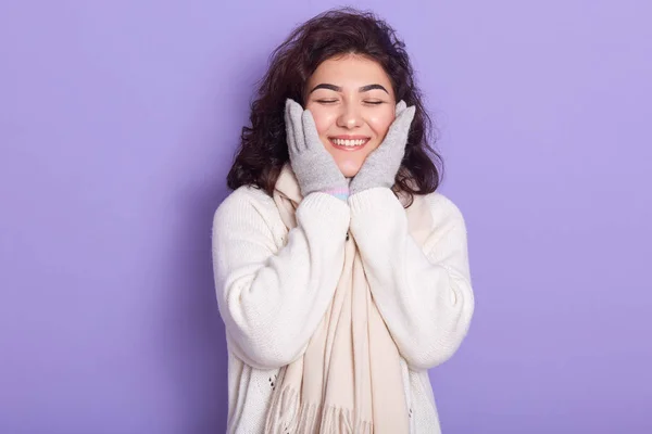 Portrait of energetic attractive female closing eyes, smiling, putting hands on cheecks, standing isolated over lilac background in studio, wearing white sweater and scarf, gloves. Emotions concept. — 스톡 사진