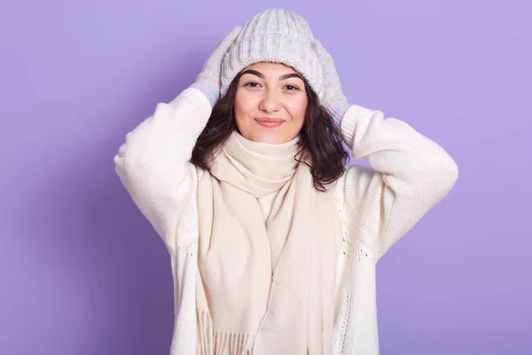 Imagen de la hermosa y agradable jovencita de pelo rizado poniendo sus manos en la cabeza, con expresión facial positiva, mirando directamente a la cámara, usando suéter blanco y bufanda, sombrero gris y guantes . —  Fotos de Stock