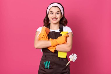Beyaz kadın lastik eldiven, önlük ve saç bandı takıyor, deterjan taşıyor, evi temizlemeye hazır, kameraya bakıyor, pembe stüdyo arka planında izole edilmiş poz veriyor. Ev işi kavramı.
