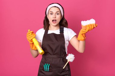 Beyaz günlük tişört, kahverengi önlük ve saç bandı takan, elinde sünger ve deterjan tutan, ağzı açık poz veren genç ev kadınının fotoğrafı, şaşırmış ve şaşırmış görünüyor, pembe duvarın üzerinde izole edilmiş.
