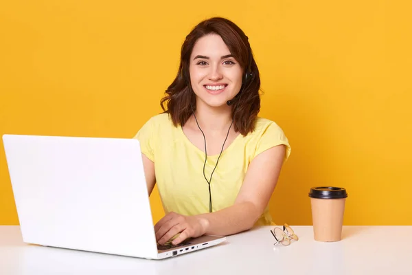 Gadis yang duduk di meja dan bekerja melalui laptop online, freelance, berbicara dengan klien, minum kopi dari cangkir kertas dan tersenyum gembira, menatap kamera dan berpose puas atas dinding studio kuning . — Stok Foto
