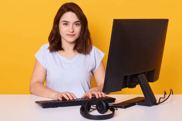 Στούντιο πλάνο της νεαρής μελαχρινής γυναίκας κάθεται στο τραπέζι μπροστά από τον υπολογιστή, ελκυστική γυναίκα ελεύθερος επαγγελματίας έχει online εργασία, πληκτρολογώντας κάτι, κοιτάζοντας άμεσα κάμερα που απομονώνονται σε κίτρινο τοίχο στούντιο. — Φωτογραφία Αρχείου