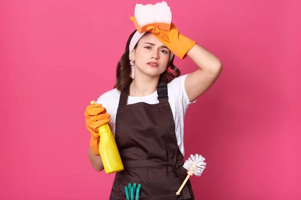 Image of upset tired housewife dresses brown apron with plunger and pins in pocket, posing isolated over rosy background and keeping hand with sponge on forehead. Cleaning and housekeeping concept.