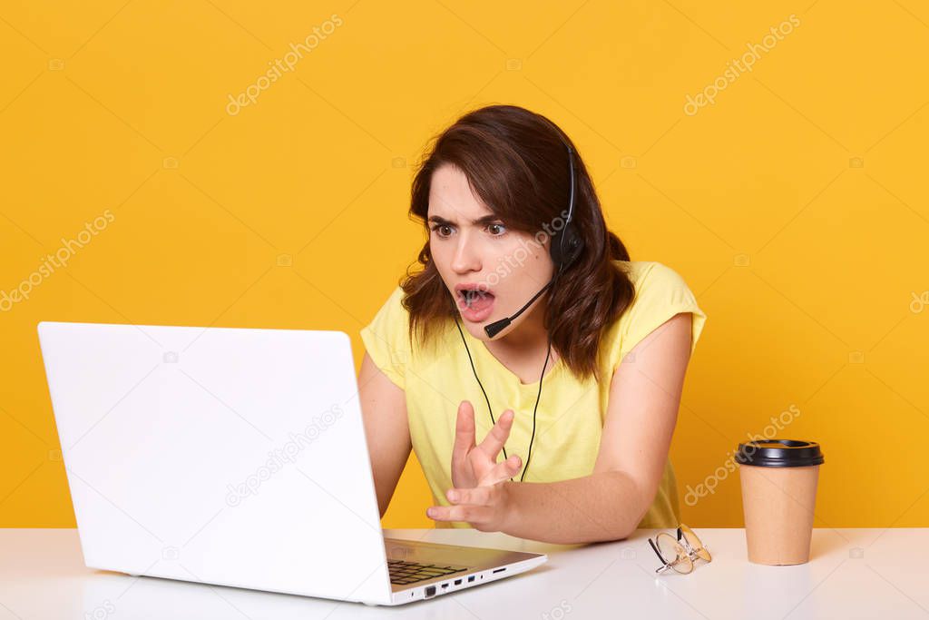 Woman seatting at table and looking at her laptop screen in consternation and horror, keeping mouth opened, wearinfcasual tshirt, posing isolated over yellow studio background, working online.