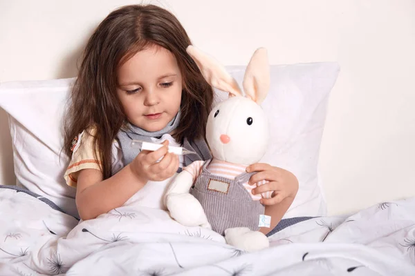 Meisje liggend in bed en het meten van de temperatuur van zachte horen met thermometer, kind liggend in bed op kussen met haar favoriete speelgoed, het dragen van grijze sjaal rond de nek, lijden aan rook, cathing koud. — Stockfoto