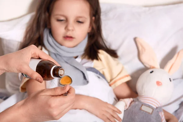 Οριζόντια φωτογραφία άγνωστου χέρι ρίχνει υγρό στο κουτάλι από το μικρό μπουκάλι με σιρόπι, πρόσωπο που νοιάζεται για το παιδί που βρίσκεται στο κρεβάτι με γρίπη και πονόλαιμο, είναι αναστατωμένος. Παιδιά και έννοια της υγείας. — Φωτογραφία Αρχείου