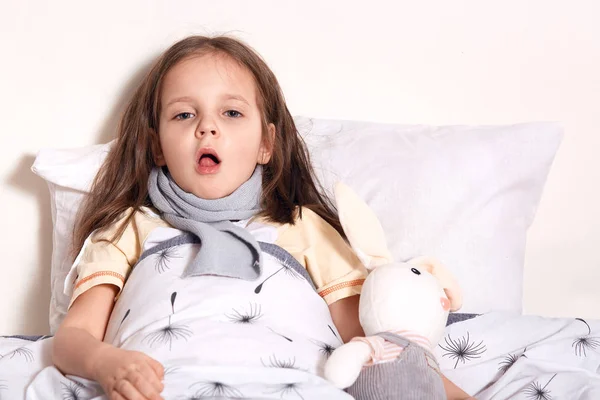 Charming cute child lying in her bed with toy, looking directly at camera, opening her mouth widely, being on sick leave, staying at home, feeling unwell, having sore throat and cough. Health concept. — Stock Photo, Image