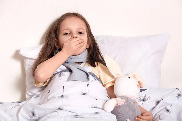 Портрет чихающего ребенка, закрывающего рот рукой, выглядящего уставшей, согревающей горло шарфом, имеющей грипп, проводящей время с игрушкой в спальне, чувствующей себя нездоровой. Концепция болезни . — стоковое фото