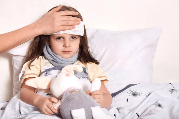 Κουρασμένο άρρωστο κοριτσάκι που έχει άγνωστο χέρι στο μέτωπο κρατώντας βρεγμένη πετσέτα για να μειώσει τον πυρετό και τον πόνο. Φτωχό βρέφος με μαντήλι γύρω από το λαιμό, που υποφέρει από πόνο στο λαιμό, κρατώντας αγαπημένο παιχνίδι. — Φωτογραφία Αρχείου