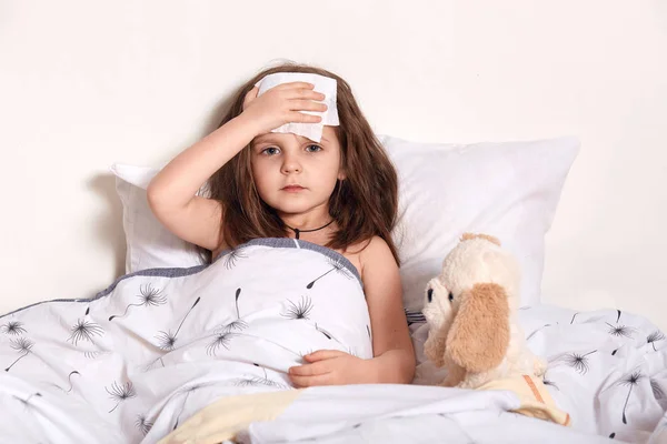 Image of bad looking child holding cold wet napkin on forehead in order to reduce fever, having caught cold, looking directly at camera, having unpleasant facial expression. Health care concept. — Stock Photo, Image