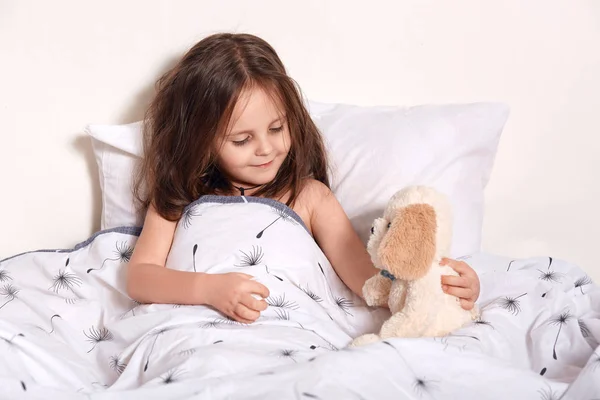 Horizontal indoor picture of cute sweet child playing in bed with her teddy bear, spending time alone in bedroom, talking with her toy, having rest on weekends. Childhood and free time concept. — Stock Photo, Image
