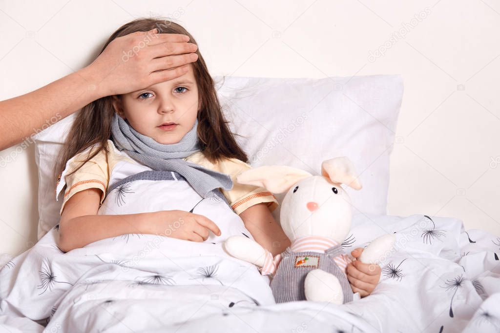 Indoor shot of little girl with fair hair lying in her bed, hugging favourite toy, having unknown hand on forehead, checking temperature, having scarf around neck, being on sick leave. Health concept.