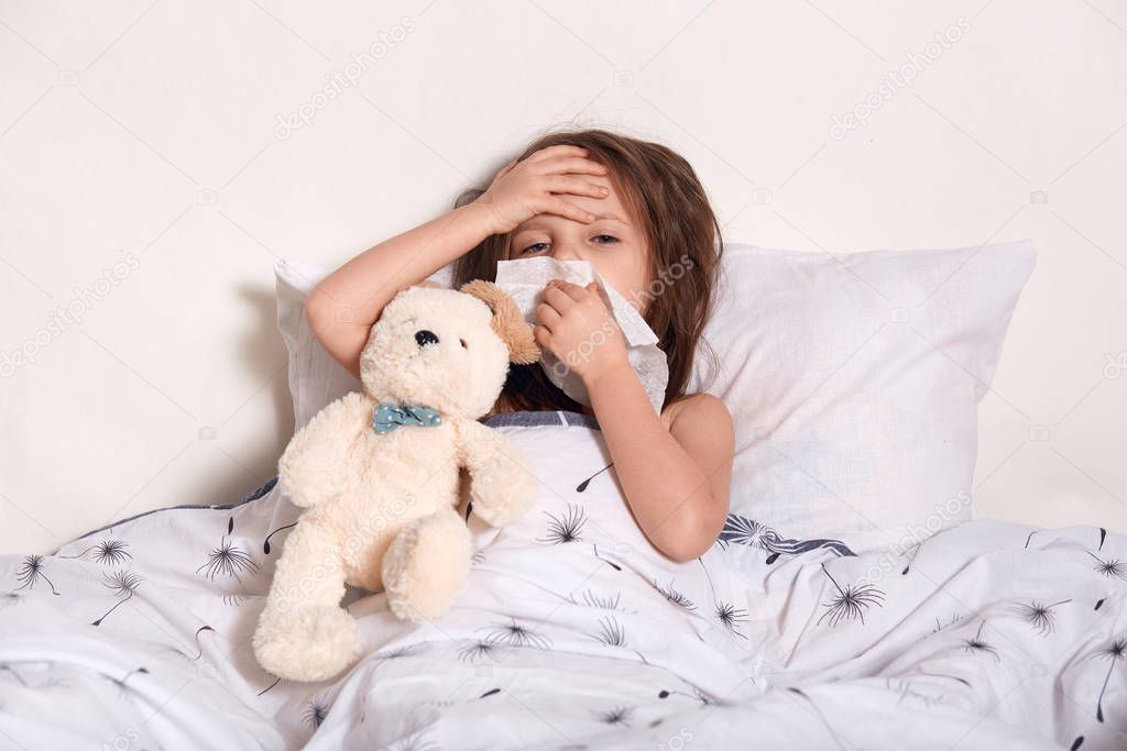 Tired ill disappointed little kid holding one hand on forehead, blowing nose in bed, using tissue, having symptoms of flu, running high temperature, having fever, suffering from pain. Health concept.