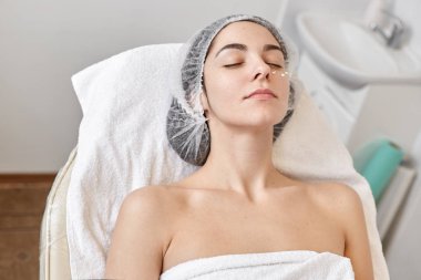 Genç, tek kullanımlık beyaz bir şapka ve havlu takan kozmetoloji dolabındaki kanepeye uzanmış, gözleri kapalı ve çıplak omuzlarıyla poz veren güzel bir kadın. Kozmetik ve güzellik kavramı.