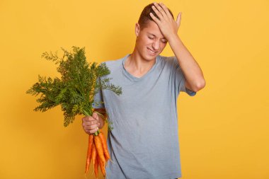 Yakışıklı delikanlının alnına eliyle dokunduğu, bir elinde havuç dolu bir demet yatay görüntü, genç adam önemli bir şeyi unuttu, stüdyoda sarı arka planda izole edilmiş bir poz verdi..