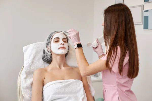 Portret pięknej kobiety układającej zawinięty biały ręcznik na wygodnej kanapie. Specjalista kosmetologii stosujący krem lub maskę do twarzy za pomocą szczotki. Piękno, spa, medycyna estetyczna i koncepcja pielęgnacji skóry. — Zdjęcie stockowe