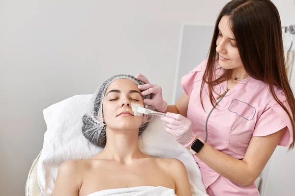 Horizontale opname van een vrouw in een kosmetologische salon, zittend op de bank, dokter cosmetoloog die reinigingsprocedures maakt, crème op het gezicht applaying. Esthetische geneeskunde, cosmetica, huidverzorgingsconcept. — Stockfoto