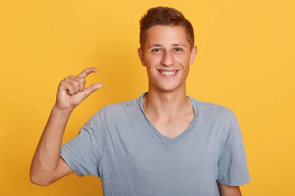 Close-up portret van knappe jongeman met stijlvolle kapsel toont kleine dingetjes. Demonstreert klein ding, poaing geïsoleerd over gele achtergrond in de studio, op zoek naar rokend naar camera. — Stockfoto