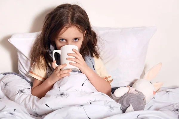 Close upportrait of child girl drinking hot tea to recover from flu, wearing pajama and scarf, dark haired child with mug in hands. Healing kids, protect immunity from seasonal virus, health concept. — Stock Photo, Image