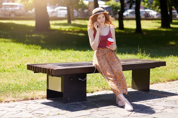 Femme chargeant son téléphone sur le chargeur de panneau solaire incorporé dans le banc assis, boire du café à emporter tout en parlant à quelqu'un via son téléphone intelligent. Technologie moderne, coconcept énergie alternative — Photo