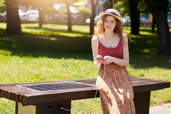 Foto av attraktiv kvinna med foxy hår, bär hatt, golvkjol och maroon t skjorta, sitter på bänk i stadsparken, ladda smarttelefon via alternativ energi. Alternativ energi, ekologi koncept. — Stockfoto