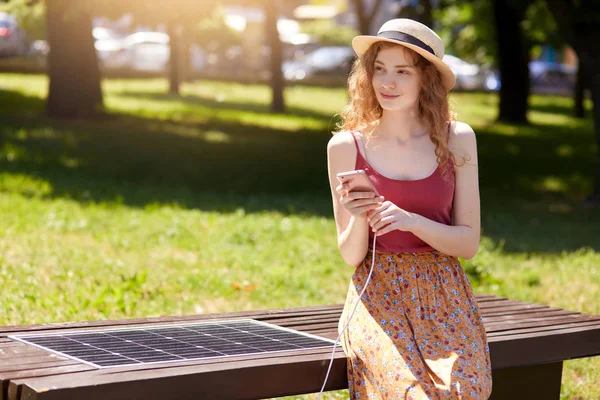 Prise de vue extérieure de téléphone de charge de femme sur le panneau solaire polyvalent gratuit, jupe féminine attrayante portant, t-shirt et chapeau, regardant de côté, semble rêveuse, en utilisant le panneau solaire dans le parc de la ville. Énergie alternative — Photo