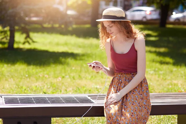 Jeune belle femme ayant du repos dans le parc, assise sur un banc innovant et chargeant un appareil mobile tout en vérifiant son réseau social, en utilisant un panneau solaire, l'écologie et le concept de technologie moderne . — Photo