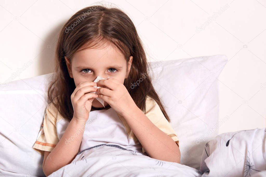 Dark haired gemale using some napkins for her running nose, looking at camera, looks sad and upset, lying in bed, catching cold, having flue, waiting for doctor pediatricion. Health care concept.