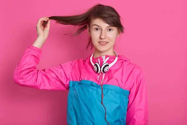 Horizontale opname van jonge behaarde donkerharige vrouw die haar bruine lange haar met opgeheven hand aanraakt terwijl ze over een roze studiomuur poseert, licht glimlacht en direct naar de camera kijkt. Mensenconcept. — Stockfoto