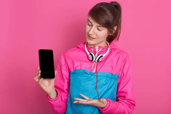 Foto close up dari wanita muda yang menarik mengenakan kemeja olahraga dan headphone di sekitar leher, menunjukkan layar smartphone kosong dan menunjuk dengan tangan, berpose terisolasi di atas latar belakang studio merah muda . — Stok Foto