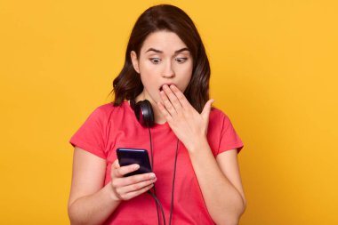Yarım boy esmer kadın portresi, tişört giyiyor, elinde akıllı bir telefon var, şaşırmış ve şaşırmış görünüyor, ağzı açık poz veriyor, sarı arka planda izole edilmiş. İnsan duyguları kavramı.
