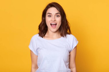 Şaşkın, genç, siyah saçlı, ayakta duran ve kameraya bakan bir kadının portresi. Şok geçiriyor, ağzını açık tutuyor, beyaz t-shirt giyiyor, keyfi yerinde. İnsan duyguları kavramı.