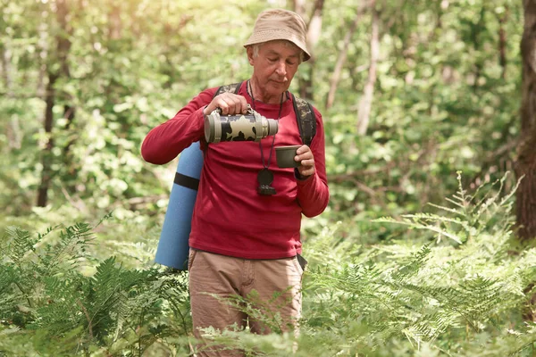 Colpo all'aperto di vecchio con borsa sul retro, tenendo thermos ed essendo pronto a drik tè caldo o caffè, avendo pigiama party, indossando cappello beige e felpa rossa, essendo appassionato di viaggi ed escursioni . — Foto Stock